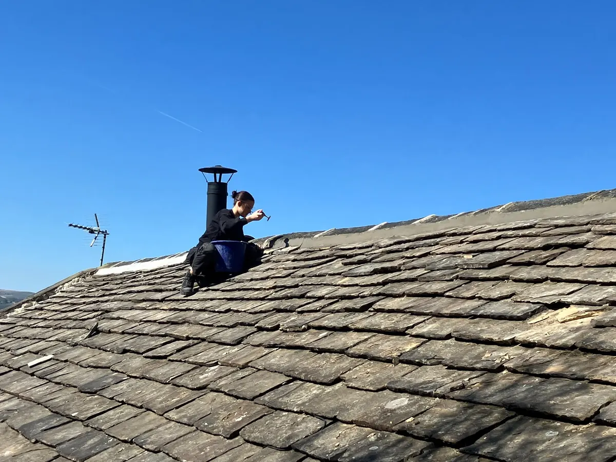 Roofer working at a chimney stack on a stone-slate roof against a clear blue sky